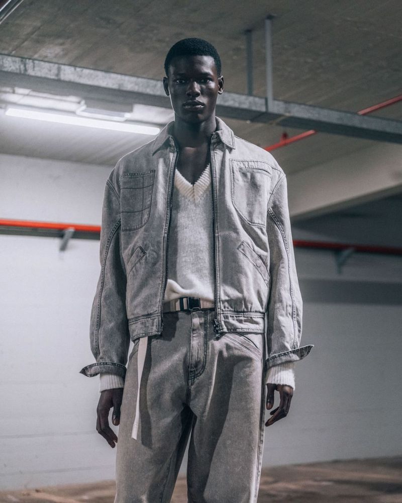 An adult man stands in a concrete environment wearing a light grey denim jacket, v-neck sweater, light grey denim jeans and a belt.