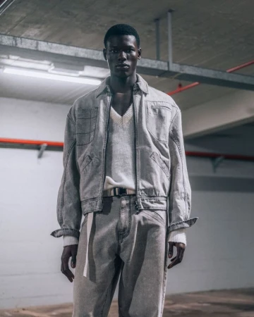 An adult man stands in a concrete environment wearing a light grey denim jacket, v-neck sweater, light grey denim jeans and a belt.