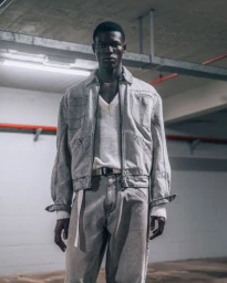 A Black male model wearing a grey jacket and pants standing in a concrete parking garage for Ferrari campaign.