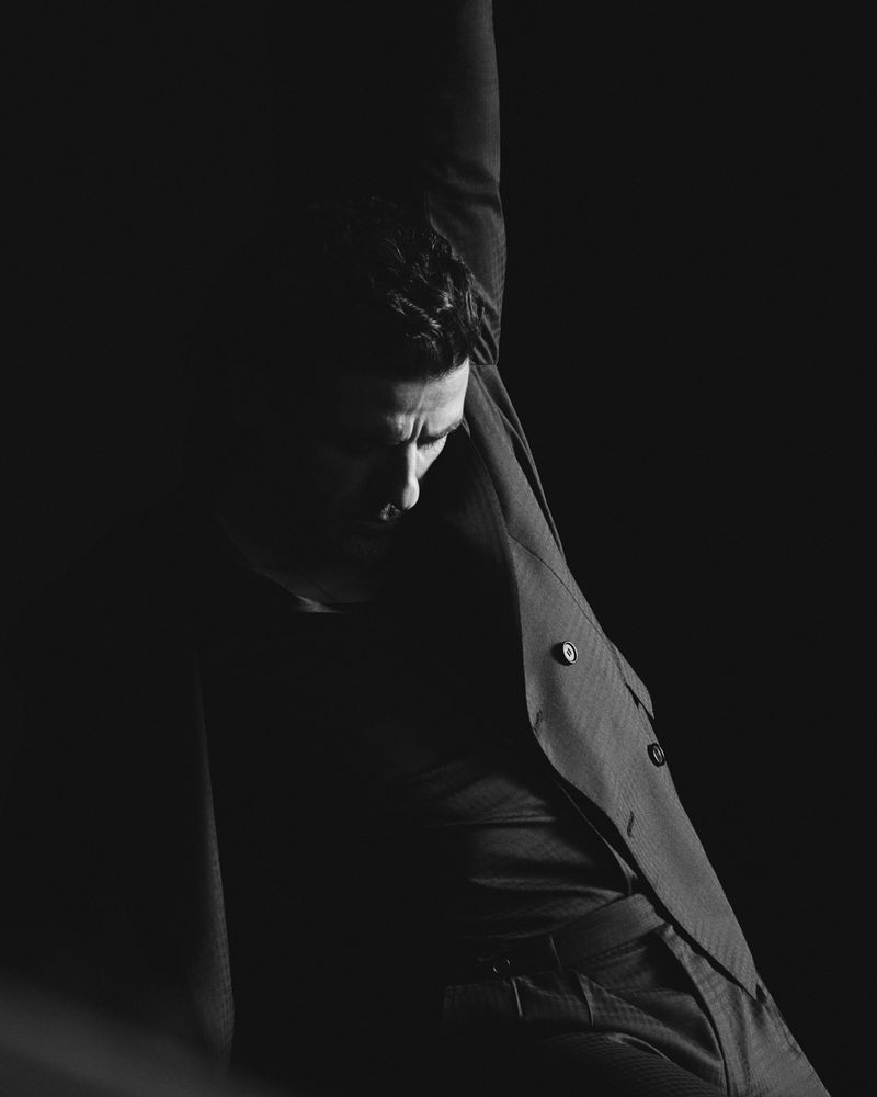 Monochrome portrait of a person in a suit with arm raised in a dark photo studio. The lighting is dramatic, emphasizing the form.