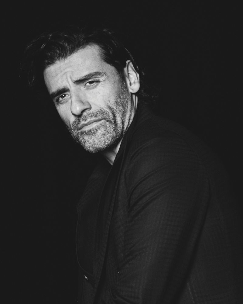 Black-and-white portrait of a man with dark hair and beard, wearing a suit jacket against a dark background. Moody and expressive.