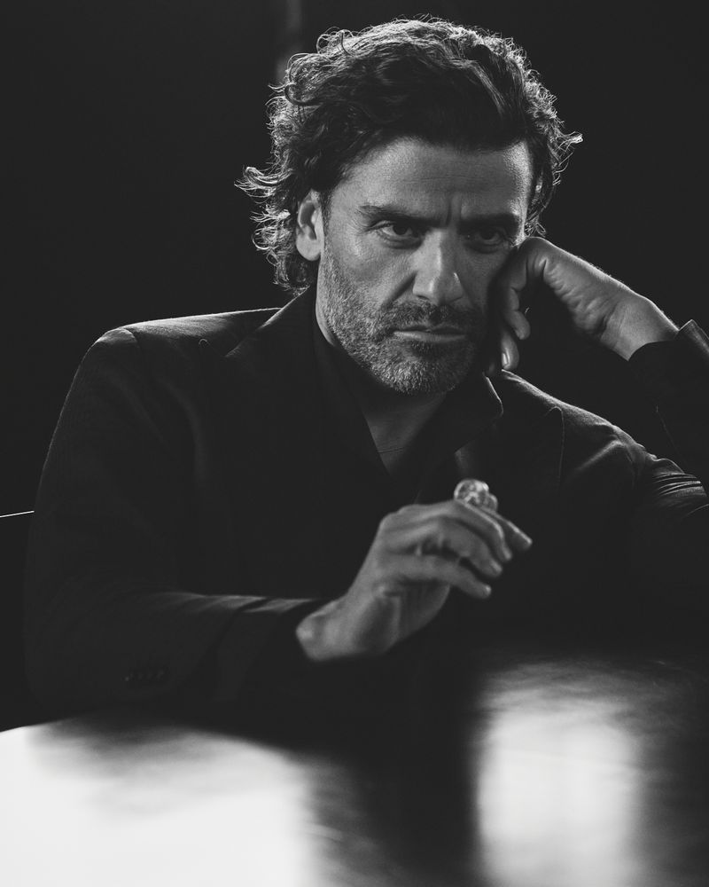 Black and white portrait of a man with dark, curly hair wearing a dark suit, posing indoors, with a hand near his face.
