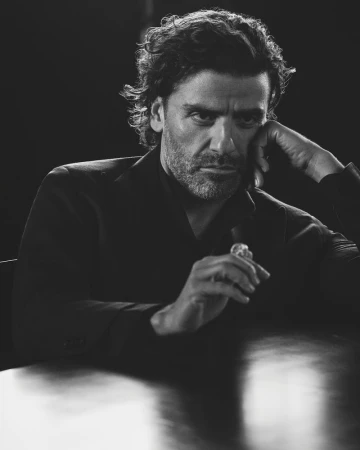Black and white portrait of a man with dark, curly hair wearing a dark suit, posing indoors, with a hand near his face.