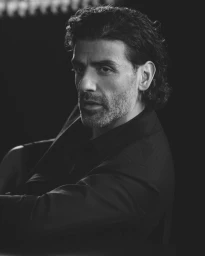 Portrait of actor Oscar Isaac wearing a dark suit against a dark background in a monochrome style campaign for Brioni.