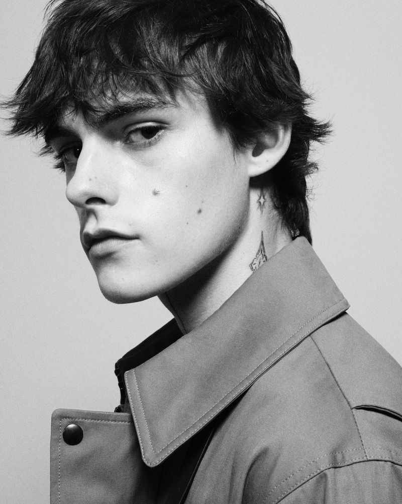 Black-and-white portrait of a young adult male with dark, tousled hair, wearing a trench coat, in a photo studio against a neutral background.