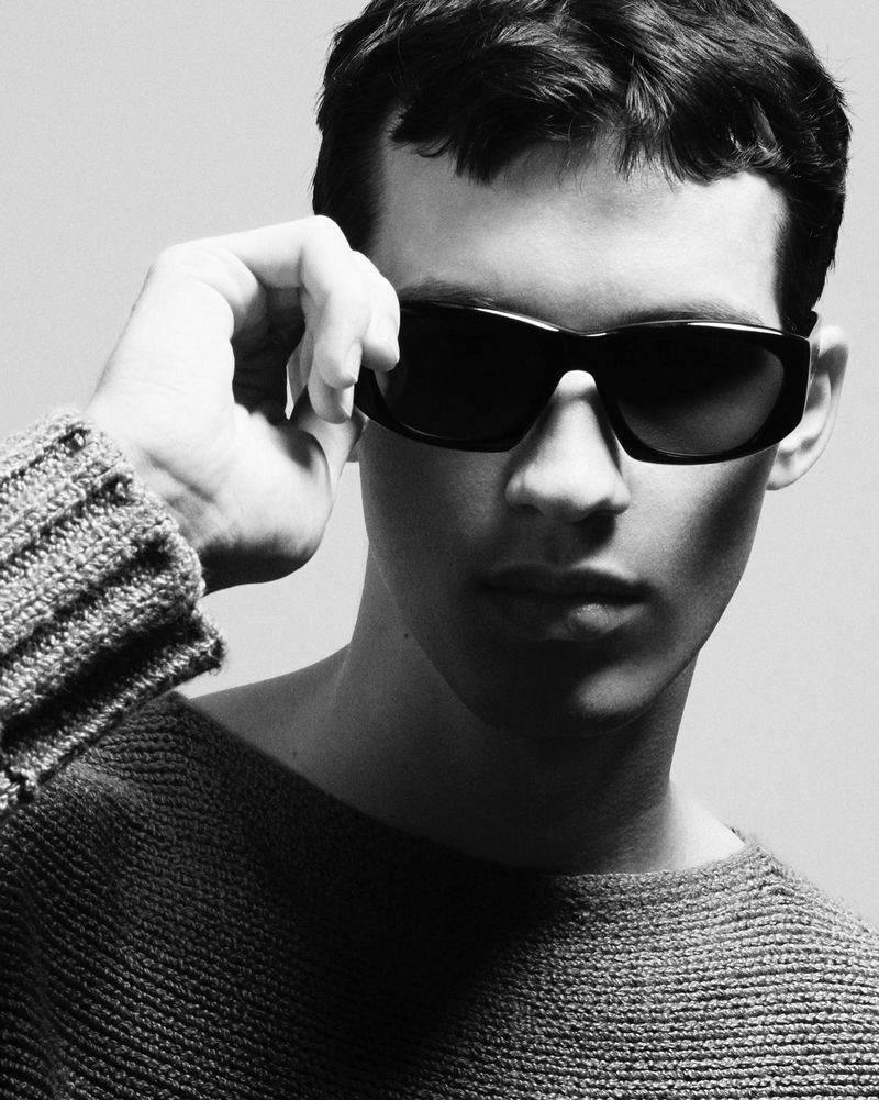 Close-up portrait of a male model wearing dark sunglasses and a textured grey knit sweater against a plain light background.