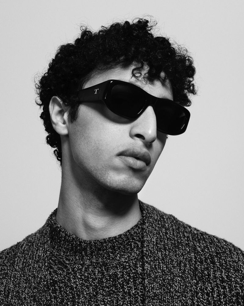 Headshot of a young adult male with curly hair wearing black sunglasses and a dark, textured knit sweater against a light background.