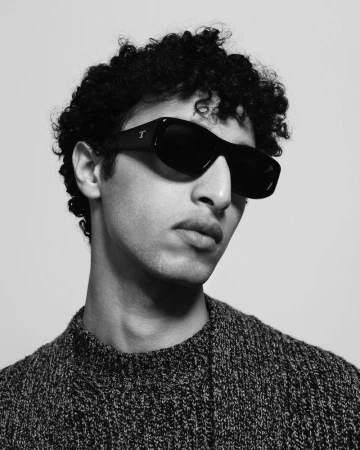 Headshot of a young adult male with curly hair wearing black sunglasses and a dark, textured knit sweater against a light background.