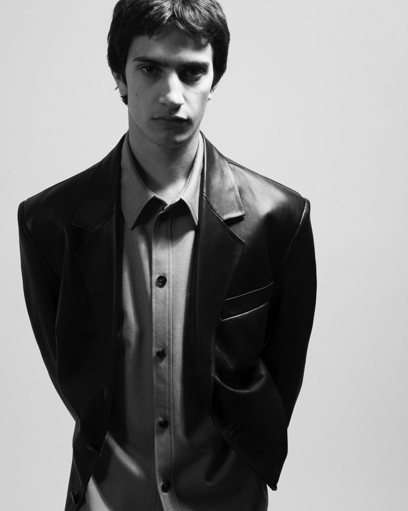 Black and white portrait of a young adult male with dark hair wearing a dark blazer over a buttoned shirt, standing against a plain background.