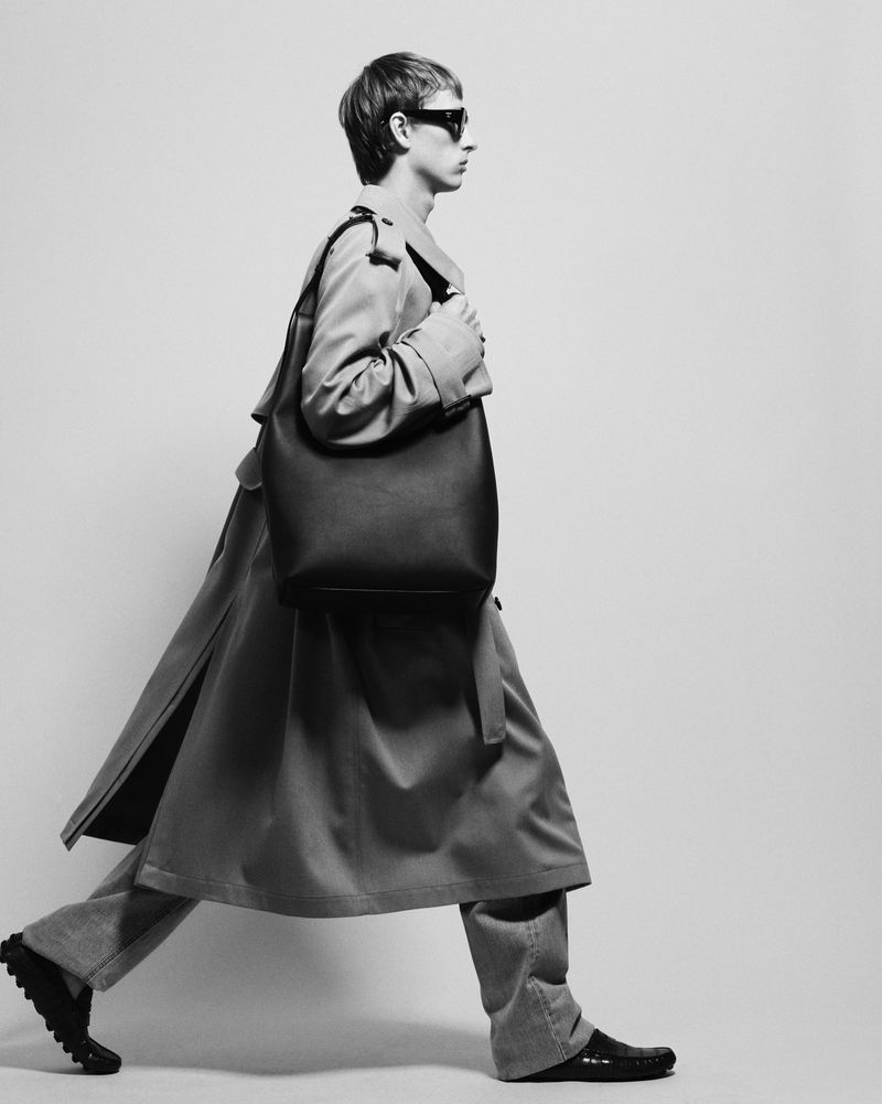 Model in profile wearing a trench coat and carrying a large shoulder bag against a light studio backdrop