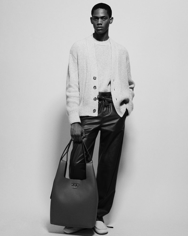 Black and white studio shot of a male model in a cardigan, sweater, and leather pants, carrying a large leather tote bag.
