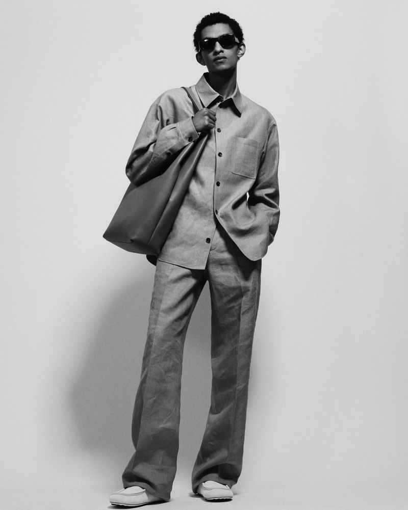 Model in dark sunglasses and matching button-down shirt and trousers carrying a large shoulder bag against a neutral background.
