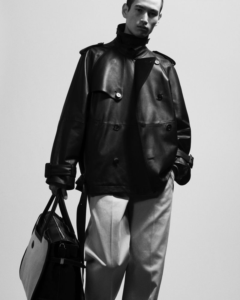 A stylish male model in a black leather trench coat and grey pants poses with a black leather duffel bag against a neutral backdrop.