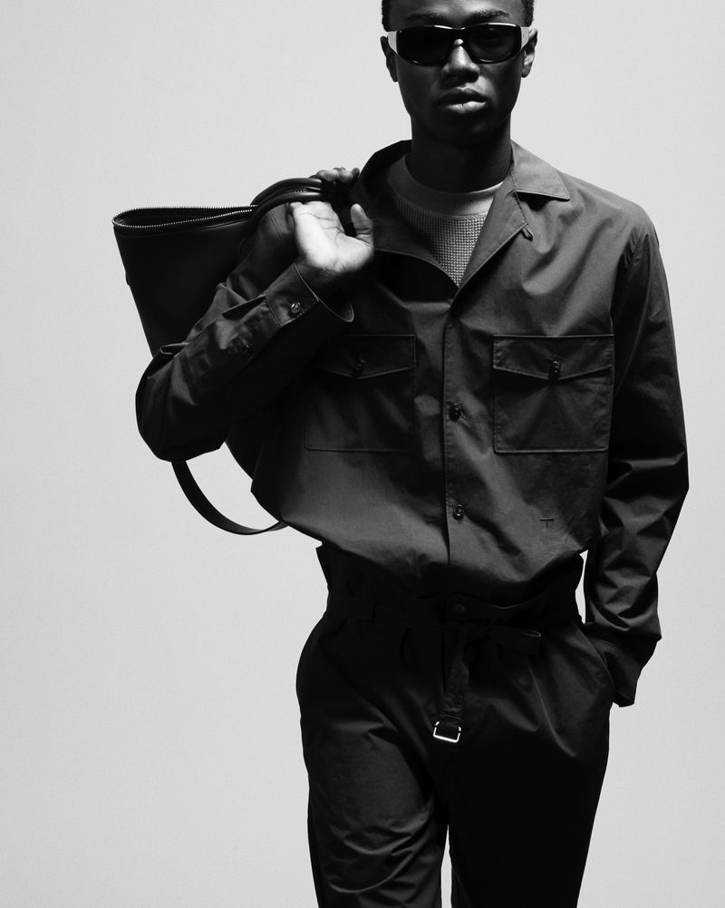 Black and white portrait of African American male model wearing a matching shirt and pants, sunglasses, and carrying a tote bag.