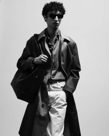 Monochrome fashion portrait of a model wearing dark sunglasses, a trench coat, and carrying a large leather shoulder bag
