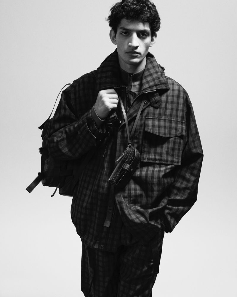 Monochrome portrait of a male model in plaid jacket, pants, backpack, and shoulder bag against a white background in a studio.