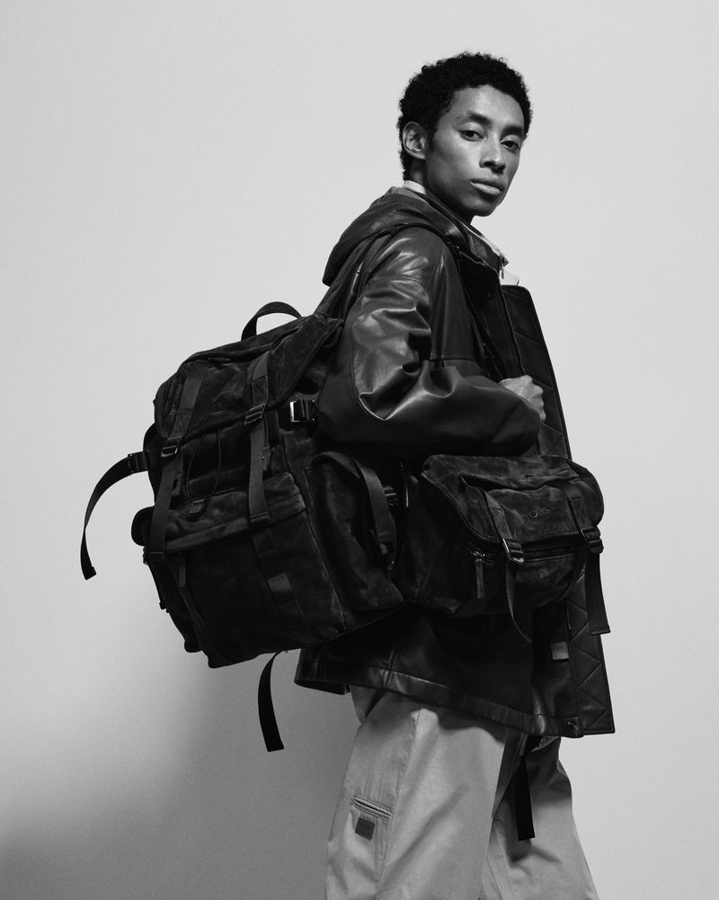 Black and white studio shot of a dark-haired man wearing a leather jacket and light pants, carrying two heavy black backpacks.