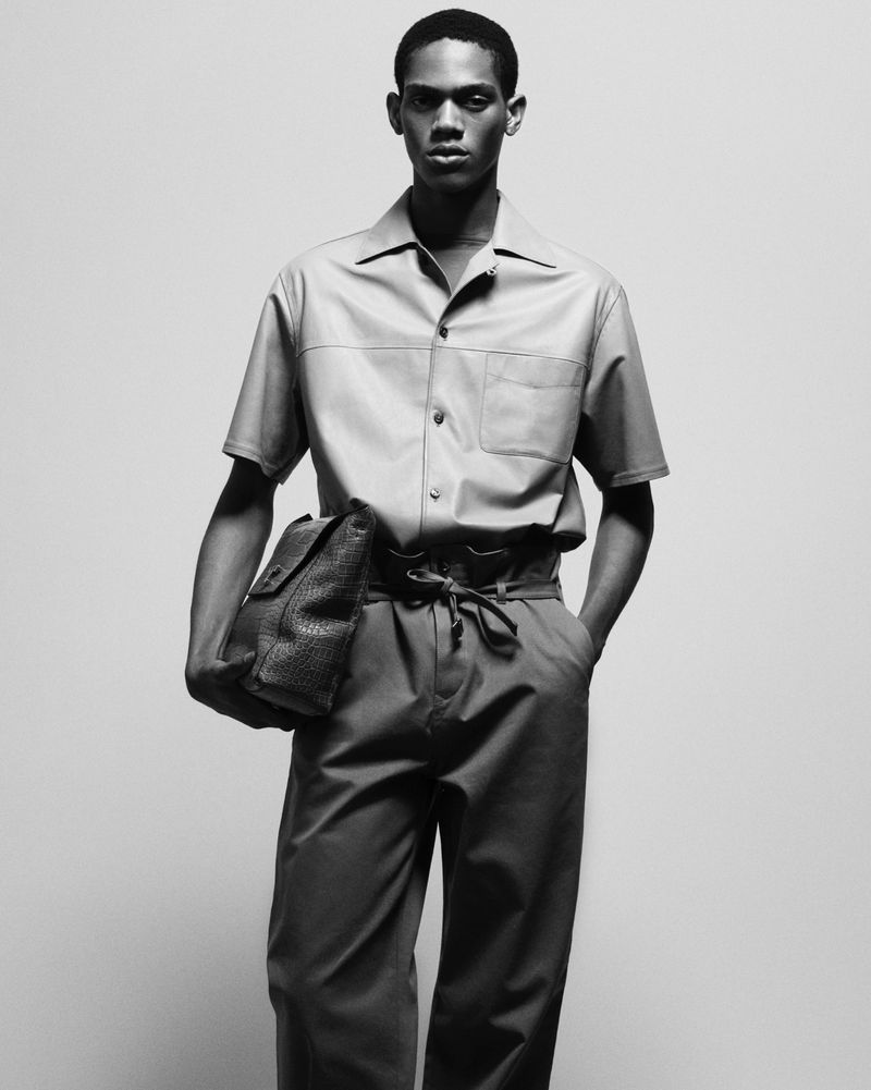 Monochrome shot of a Black male model in casual shirt and trousers, holding a leather satchel against a neutral backdrop.