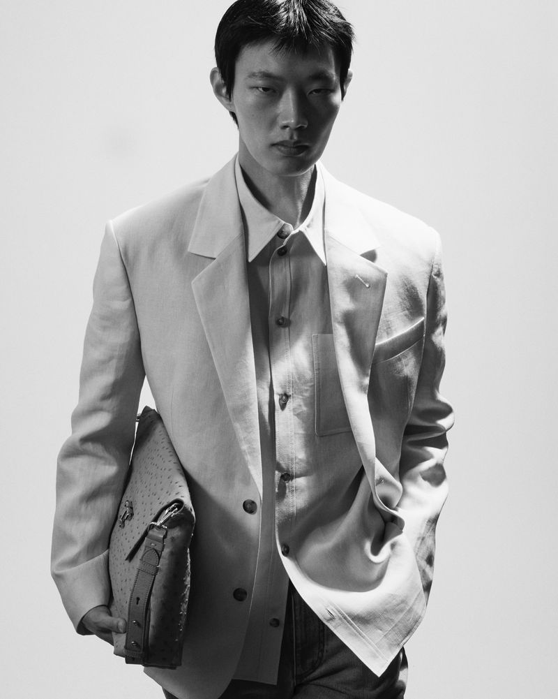 A young Asian man in a linen suit stands against a bright background, holding a leather bag, in a black and white fashion portrait.