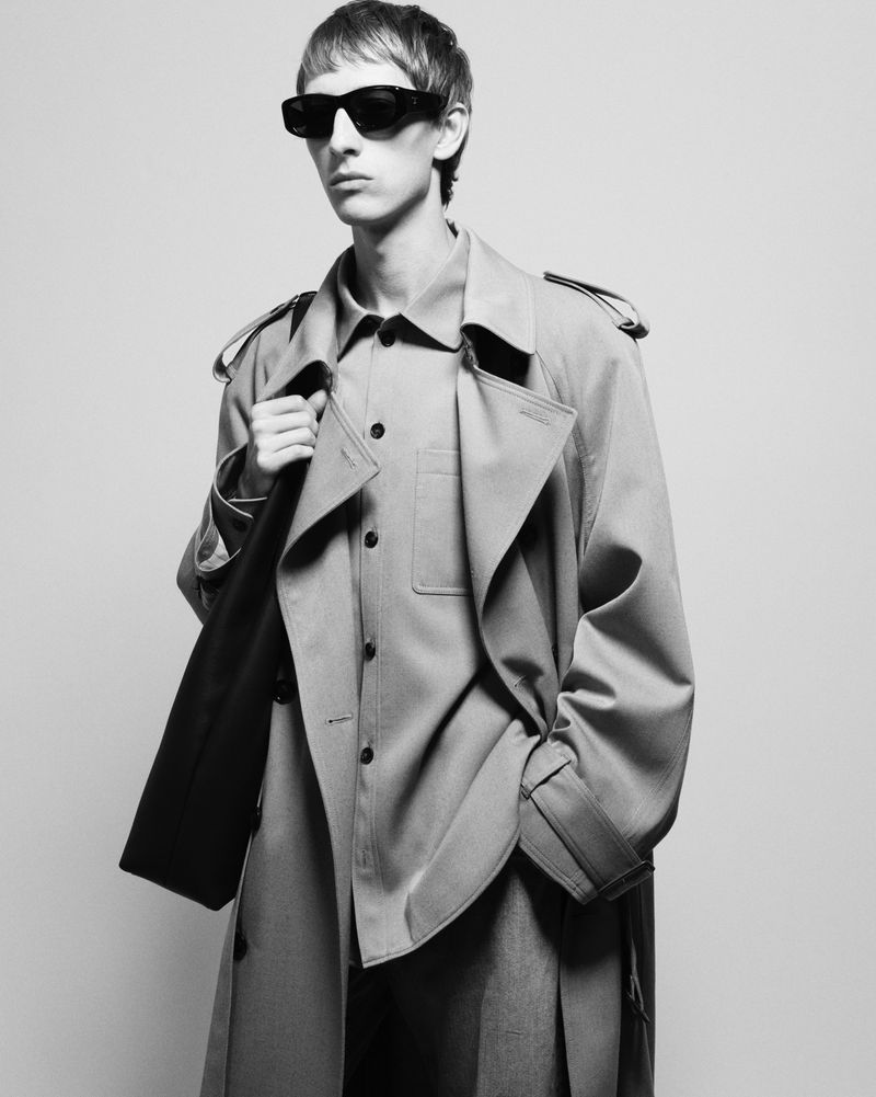 Black and white portrait of a slender male adult wearing sunglasses and a trench coat in a studio with neutral lighting.