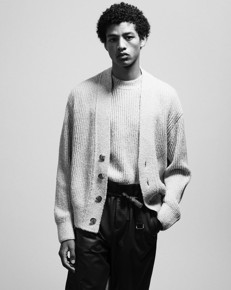 Black and white studio shot of an adult male model with curly hair wearing a knit sweater and cardigan with leather pants.