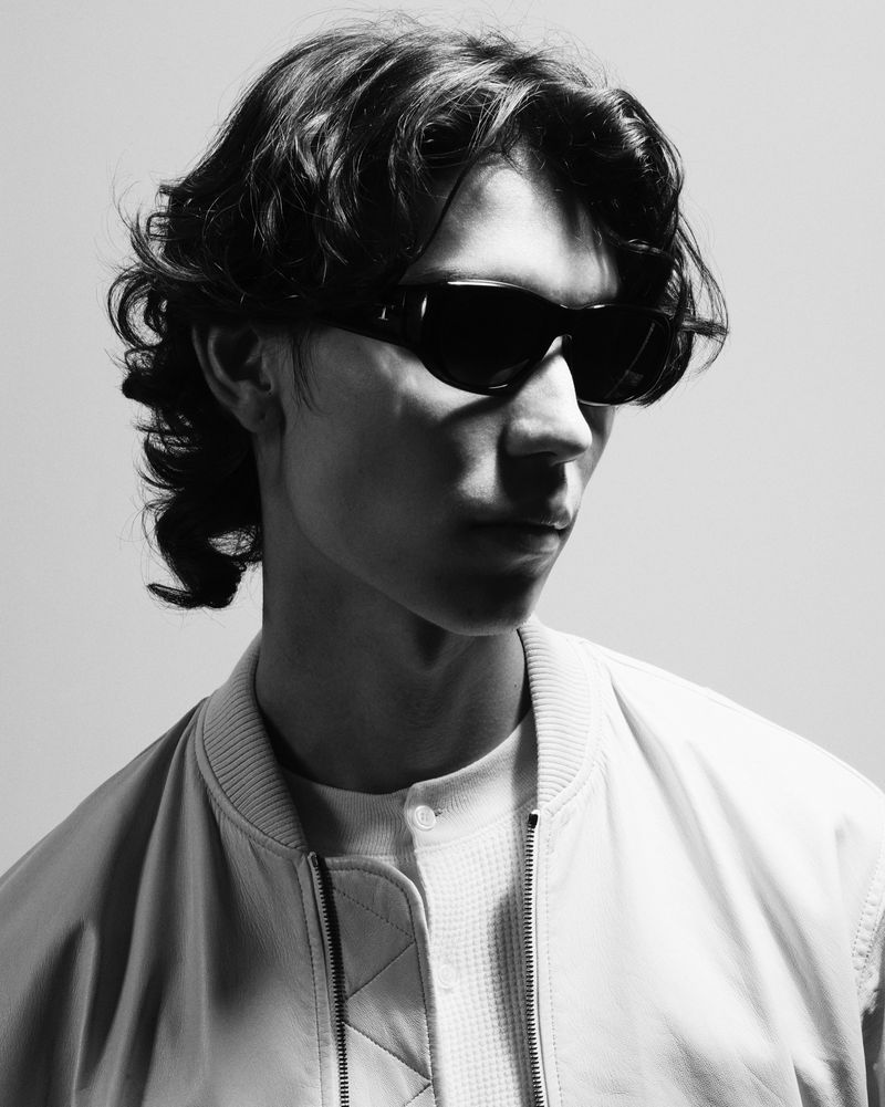 Model wearing dark sunglasses and a light-colored jacket against a plain background