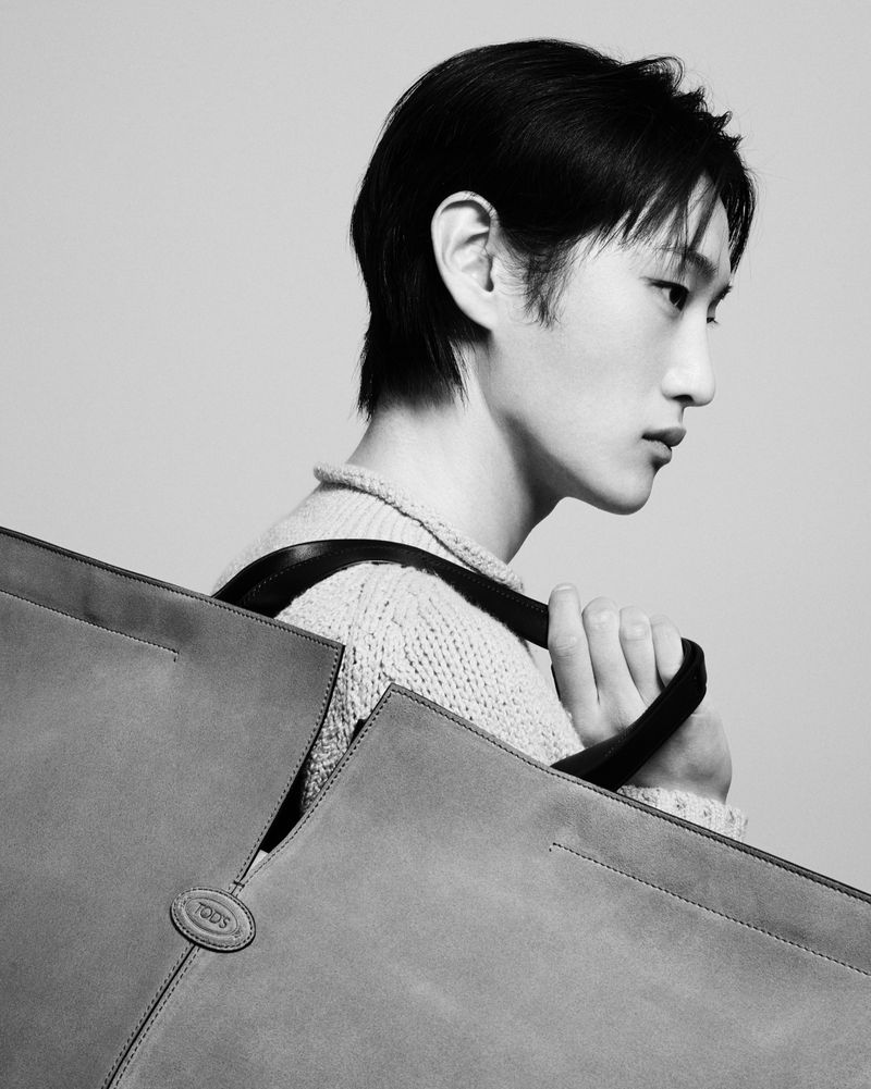 Monochrome fashion portrait of an androgynous model holding a large grey Tod's handbag in a bright neutral-toned studio setting.