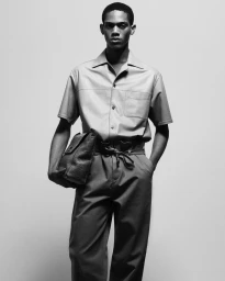 Black man standing against a seamless background wearing a button-down shirt and holding a textured leather handbag