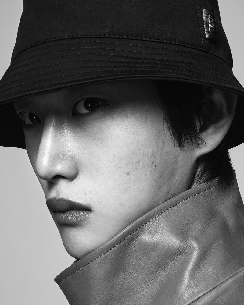 Close-up portrait of a model wearing a black bucket hat and high-collar leather coat against a grey background.