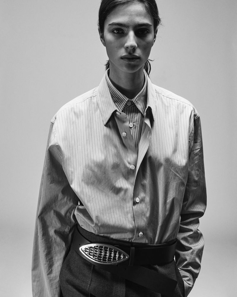 A black and white studio shot of a stylish woman posing in a striped shirt with a wide belt and an oval metal buckle.