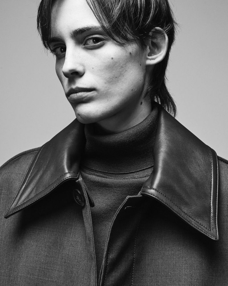 Monochrome portrait of a young adult male with dark hair wearing a turtleneck and coat with a leather collar in a studio setting.