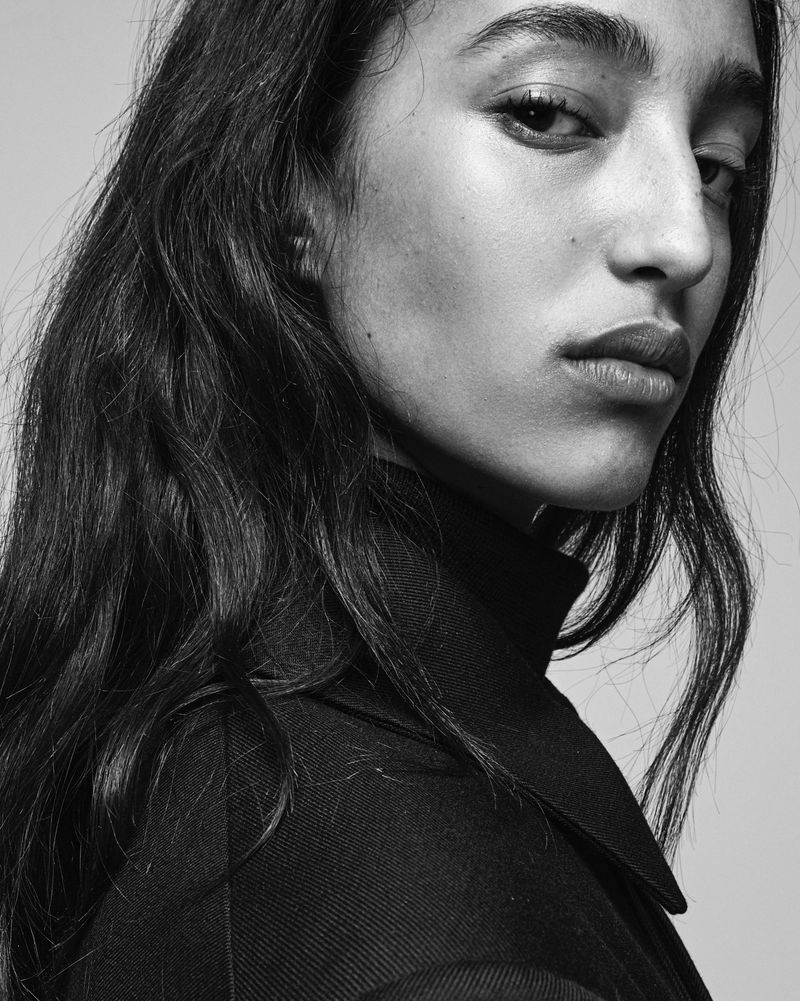 Portrait of a young woman with dark, wavy hair wearing a dark turtleneck sweater and coat, in a black and white studio shot.