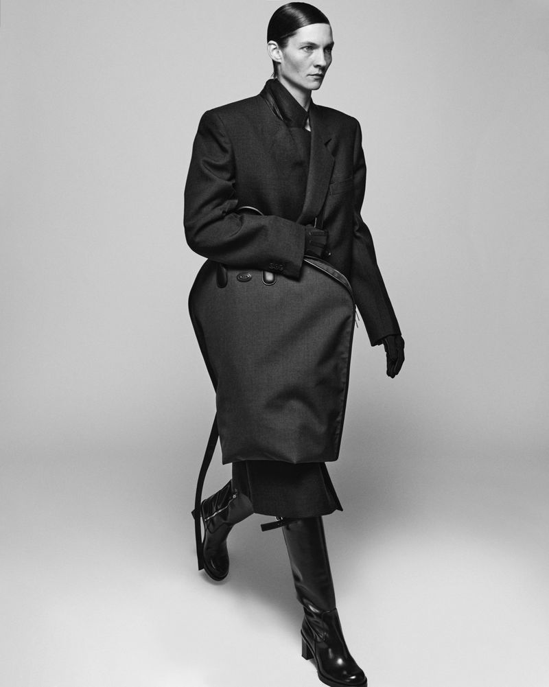 A single adult woman walks in a monochrome studio shot wearing a dark jacket, skirt, gloves and high boots against a light background.