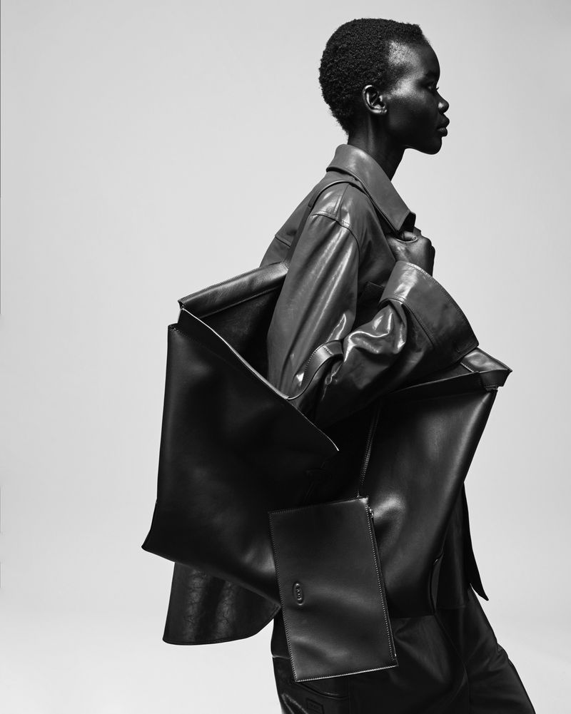 Black and white fashion portrait of a Black woman in a leather jacket with an oversized leather bag, posing in a photo studio.