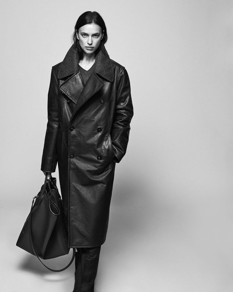 Black and white portrait of a woman in a dark leather trench coat, carrying a large handbag against a neutral studio backdrop.