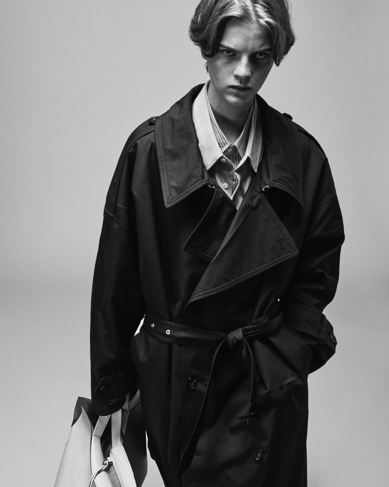 Black and white fashion shot of a young adult man wearing a trench coat and holding a bag in a neutral studio setting.
