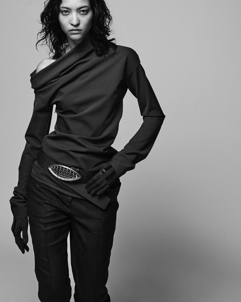 A woman in a stylish, draped top and dark pants poses in a studio, showcasing a modern, asymmetrical fashion look in black and white.