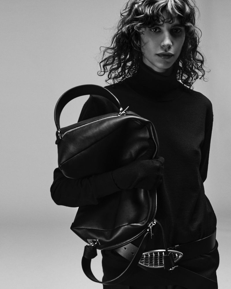 Black and white fashion shot of an adult female model holding a black leather purse with zipper details against a grey background.