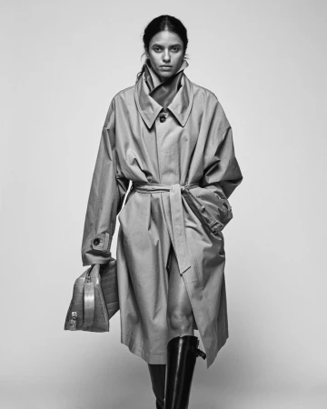 Model wearing a belted trench coat and tall boots holding a leather handbag against a plain grey studio backdrop