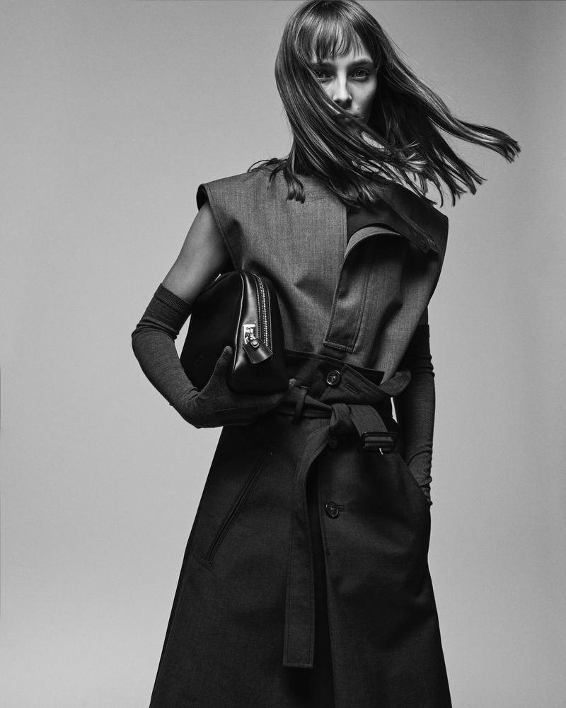 Black and white portrait of a stylish woman wearing a coat dress, holding a handbag, with a neutral studio backdrop.