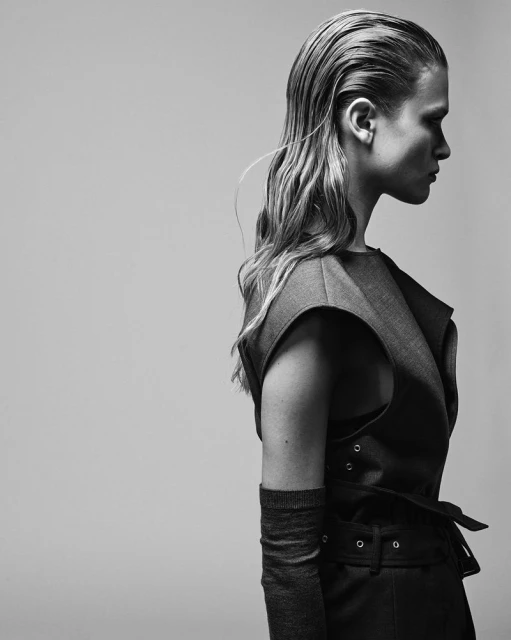 Black and white fashion portrait of a blonde adult woman in a vest and glove posing in profile on a neutral background in studio lighting.