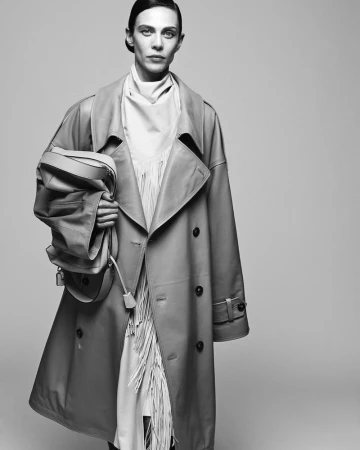 Model wearing a trench coat and holding a leather handbag against a grey seamless studio background.