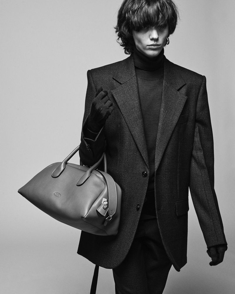 Black-and-white fashion shot: model poses in tailored blazer holding leather Tod's handbag against a neutral backdrop, studio lighting.
