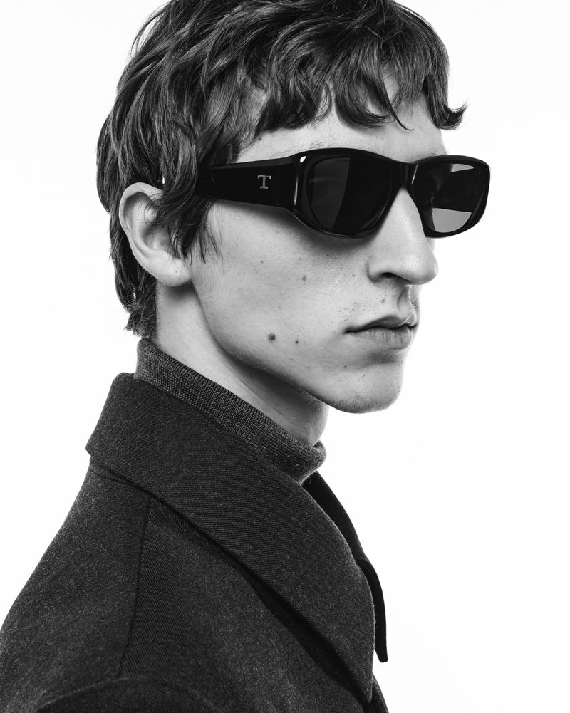 Monochrome portrait of a man wearing sunglasses and a dark-colored coat, side view against a bright white background. Fashion editorial style.