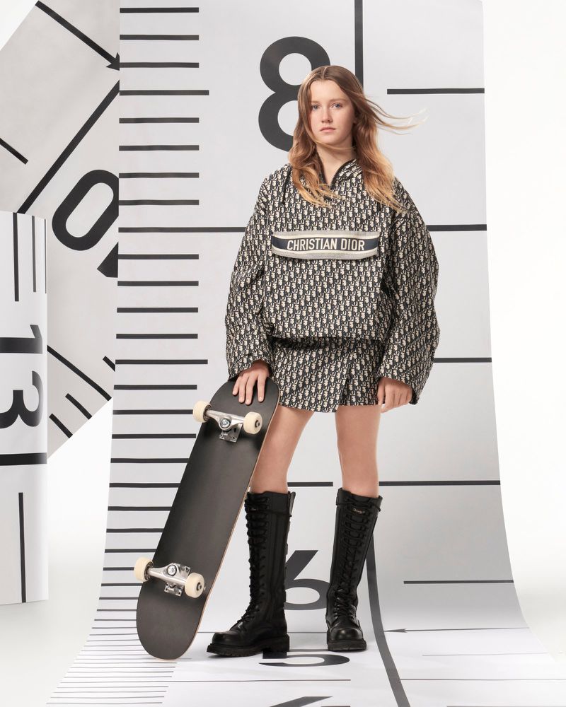 Young female model in patterned Dior outfit with skateboard, posing in a studio against a white backdrop with black ruler markings.
