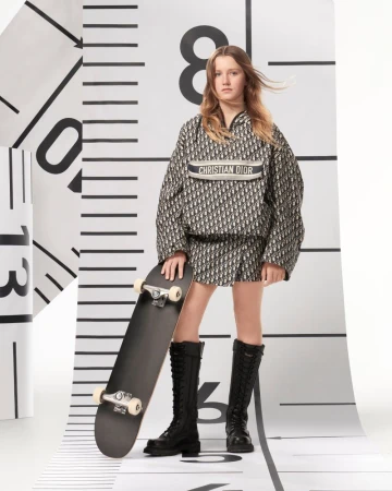 Young female model in patterned Dior outfit with skateboard, posing in a studio against a white backdrop with black ruler markings.