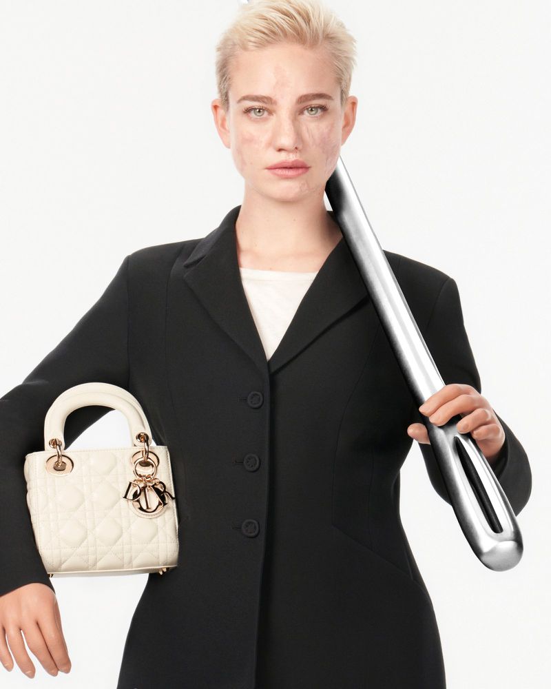 Woman in black blazer poses in studio with a white handbag and an abstract metal object, featuring short blonde hair and neutral expression.