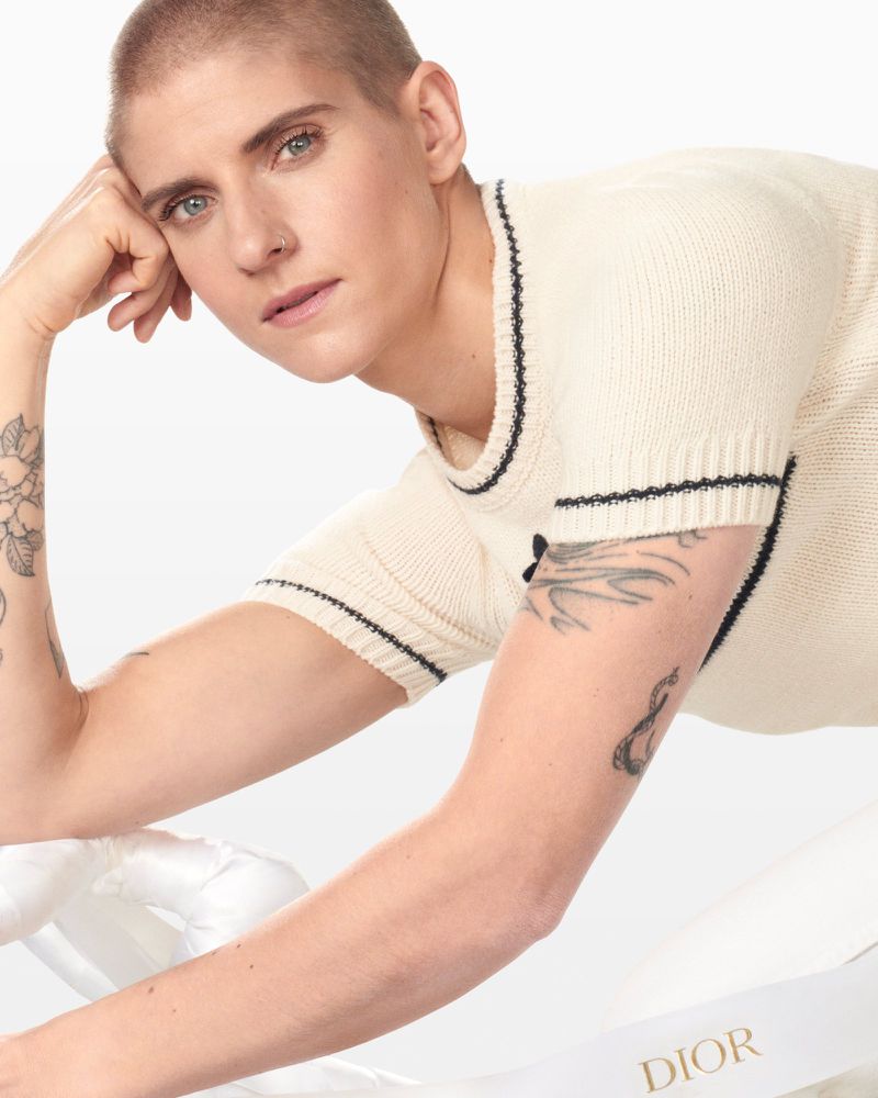 Close-up studio shot of a fair-skinned adult with a shaved head wearing a cream-colored knit shirt, Dior ribbon prop. Beauty, fashion.