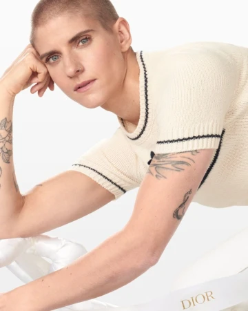 Close-up studio shot of a fair-skinned adult with a shaved head wearing a cream-colored knit shirt, Dior ribbon prop. Beauty, fashion.