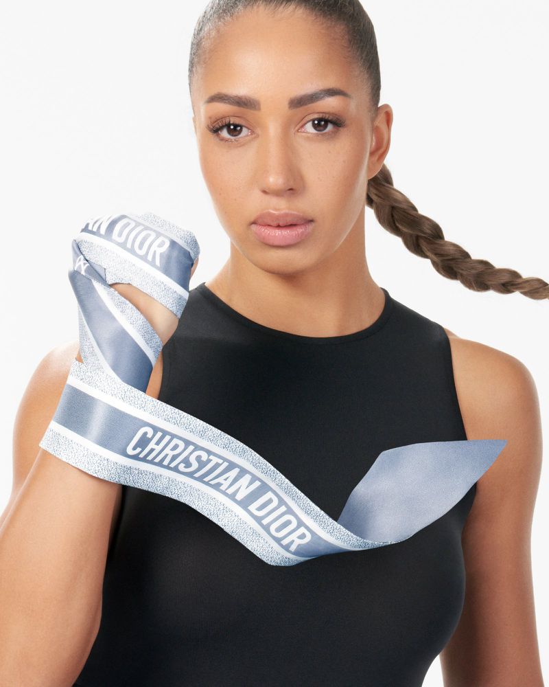 Athletic woman in workout attire holds a ribbon printed with 'Christian Dior' in a bright indoor studio environment.